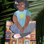 A colorful paper cutout of a smiling child with glasses, a blue shirt with a yellow star, and curly hair sits cross-legged on a quilt, placed on large green palm leaves.