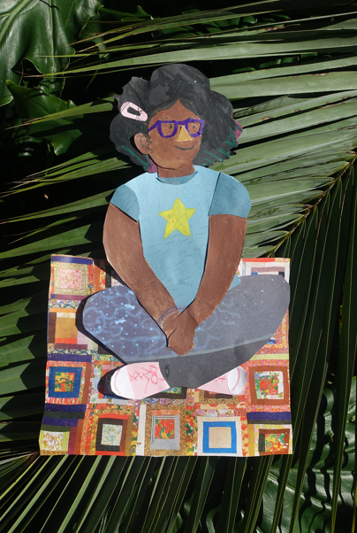 A colorful paper cutout of a smiling child with glasses, a blue shirt with a yellow star, and curly hair sits cross-legged on a quilt, placed on large green palm leaves.
