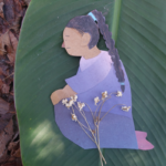 A paper cutout of a person kneeling is placed on a large green leaf, holding small dried flowers, with brown leaves scattered in the background.