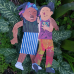 Two colorful paper cutouts of smiling children stand arm in arm on green leafy plants, one in a striped skirt and the other in patterned pants, both appearing joyful and playful outdoors.